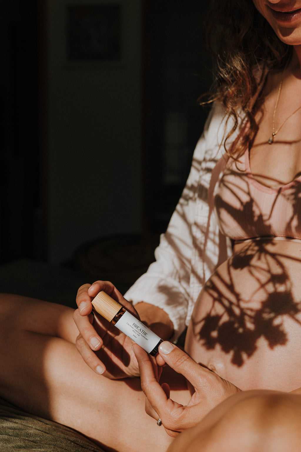 A close-up photo of a pregnant woman holding 'Breathe' birth roller blend, with warm sunlight casting leafy shadows across her belly and white top.