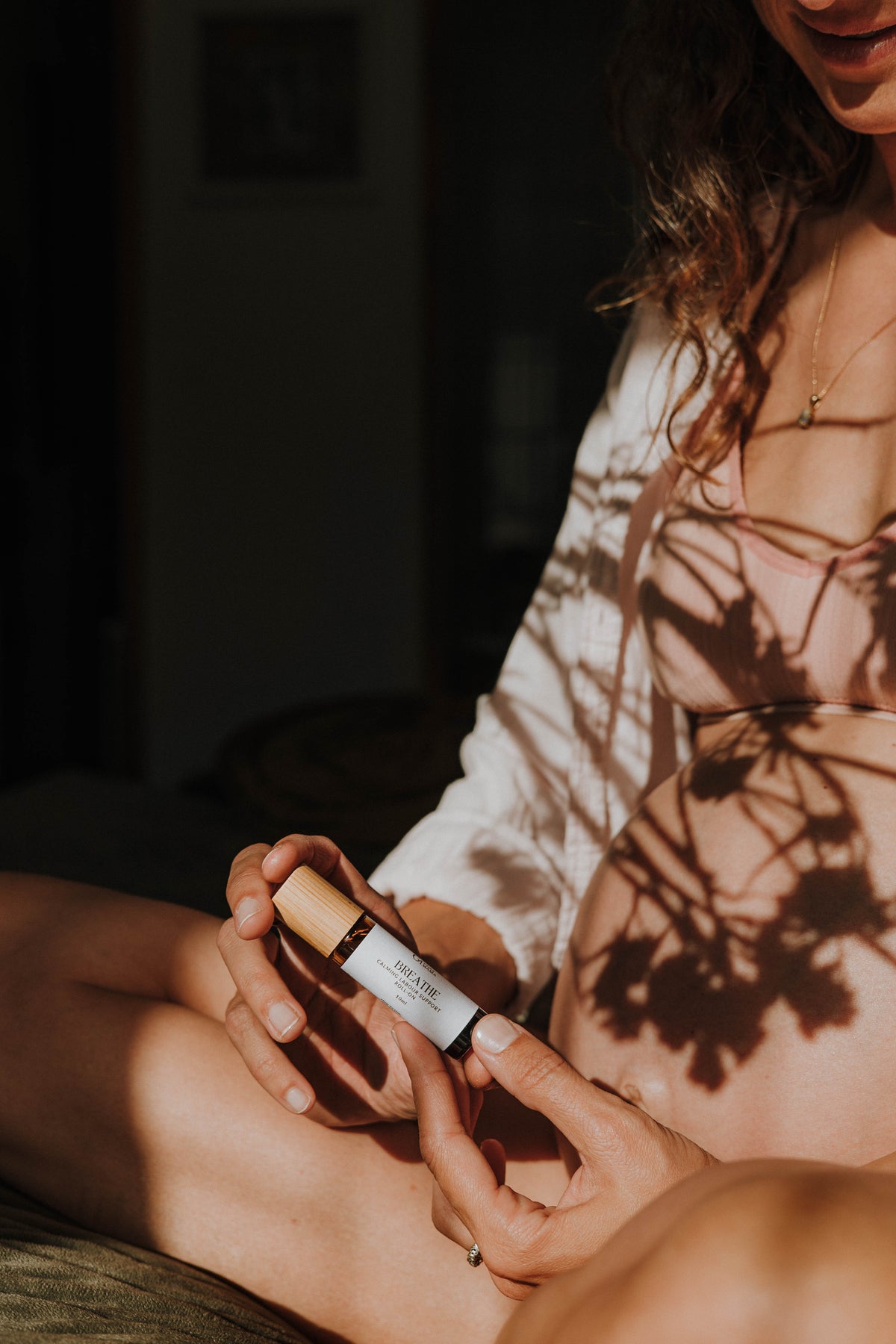 A close-up photo of a pregnant woman holding 'Breathe' birth roller blend, with warm sunlight casting leafy shadows across her belly and white top.