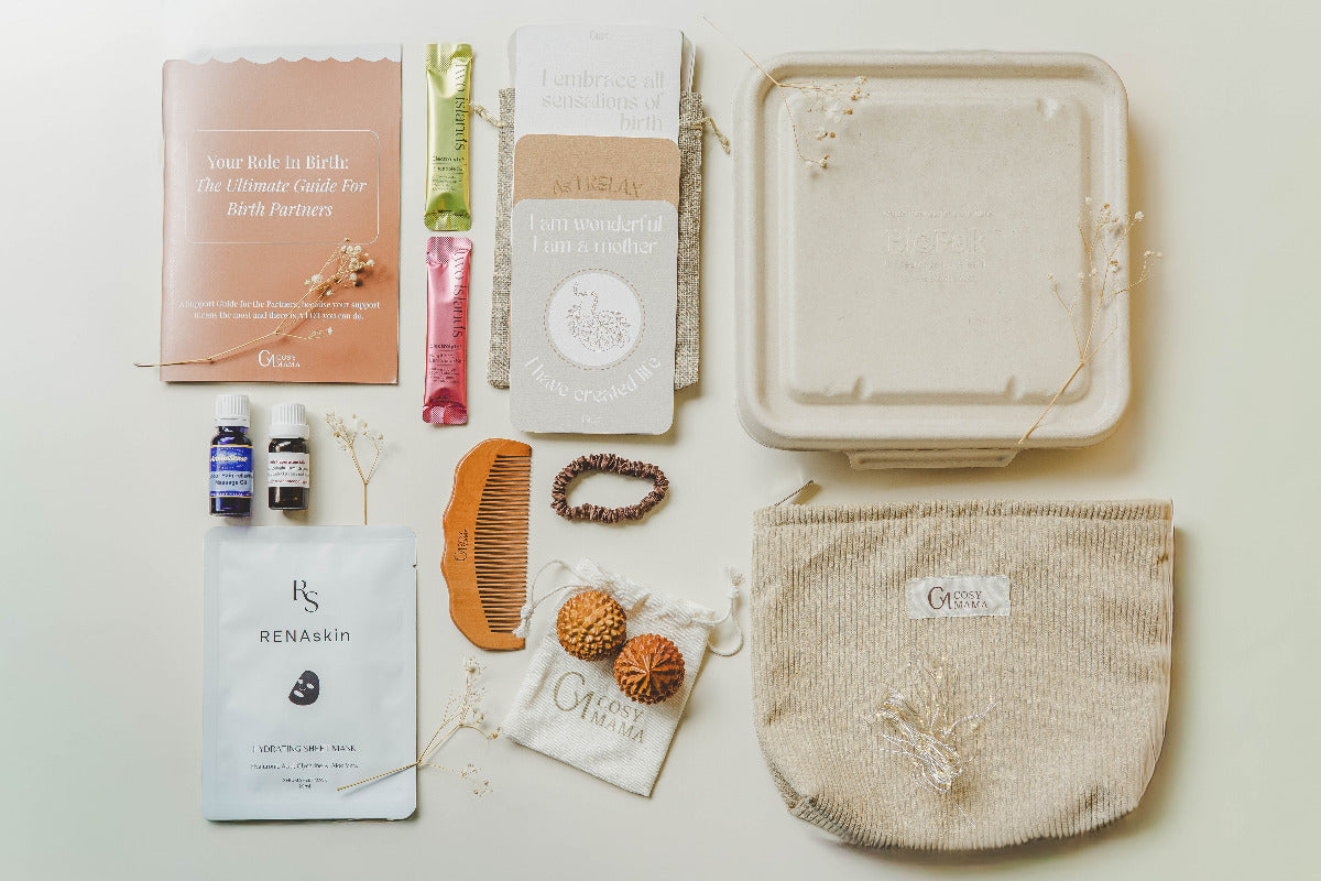 Entire Nurtured Birth Bundle from Cosy Mama on a beige background. The range includes a beige placenta container, birth affirmation cards, electrolyte sachets, birth orbs -spikey wooden balls, wooden birth comb, fairy lights, massage oil, face mask and silk hair tie.