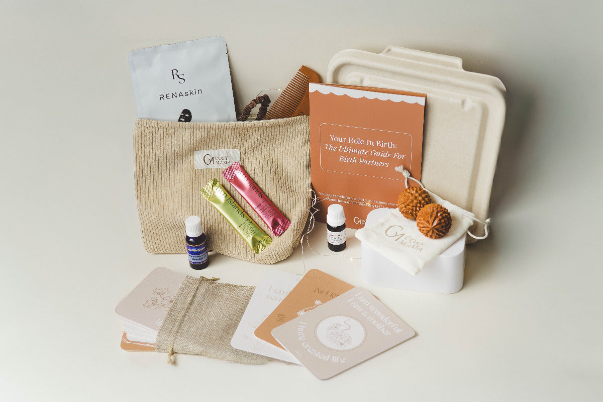 Entire Nurtured Birth Bundle from Cosy Mama on a beige background. The range includes a placenta container, birth affirmation cards, electrolyte sachets, birth orbs, birth comb, fairy lights, massage oil, face mask and silk hair tie.