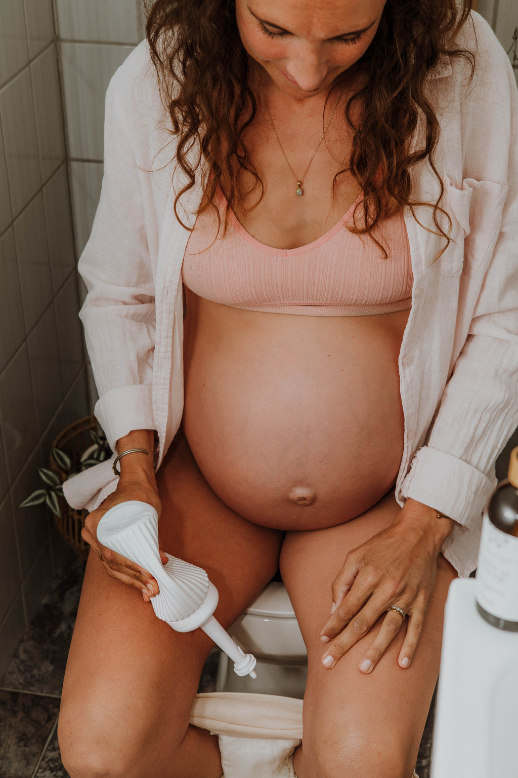 A pregnant woman sitting on a toilet in a bathroom, holding a Peri Spray Bottle. She is wearing a light pink ribbed bra and an open white button-up shirt.