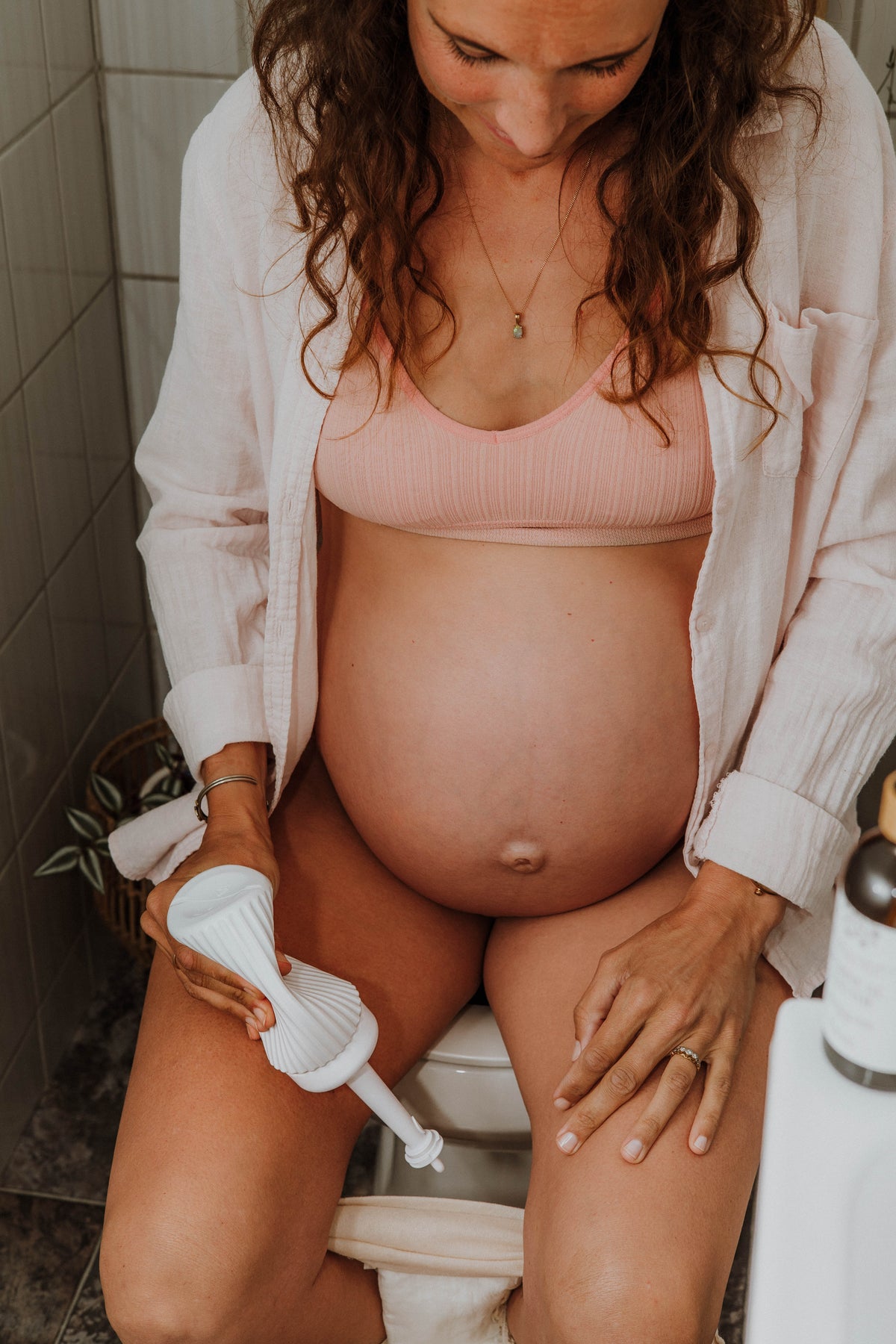 A pregnant woman sitting on a toilet in a bathroom, holding a Peri Spray Bottle. She is wearing a light pink ribbed bra and an open white button-up shirt.