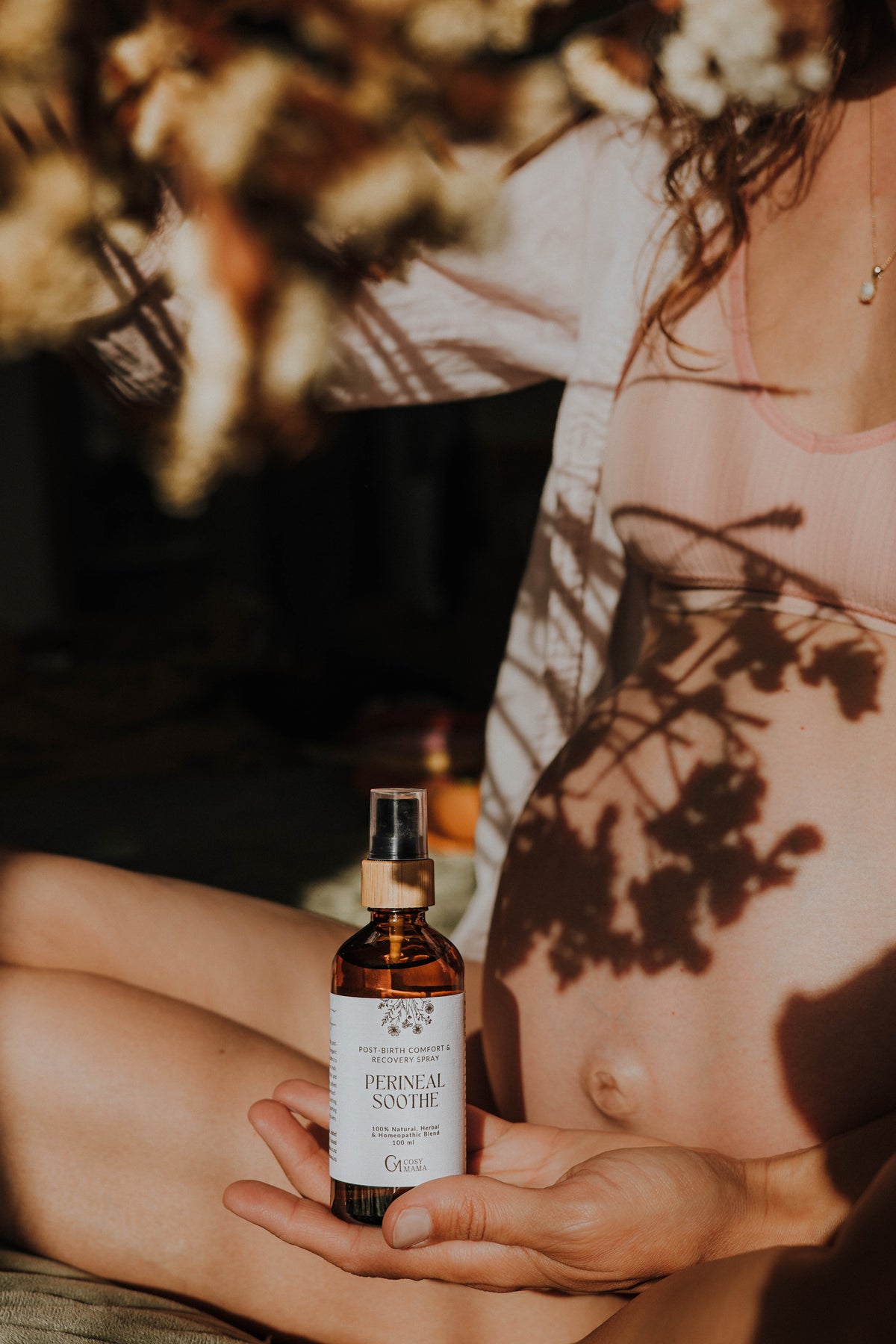 A person holds a small amber spray bottle labeled 'Perineal Soothe' in their hands, with warm sunlight and leaf shadows cast across their bare torso and lap. The scene is softly blurred with dried flowers in the foreground.