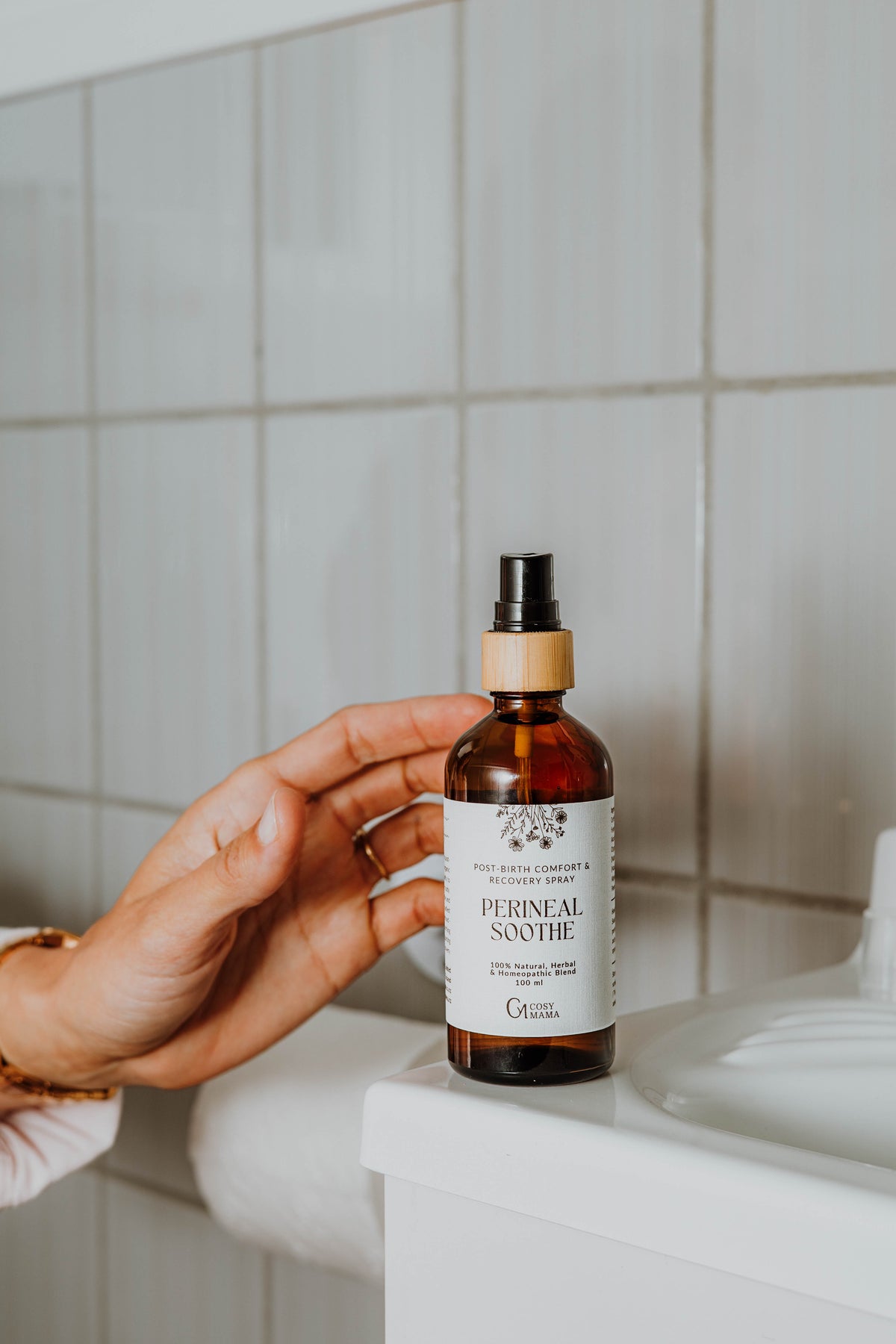 A hand holds a bottle of Perineal Soothe spray against a tiled bathroom background.