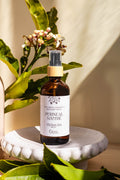 A bottle of Perineal Soothe post-birth comfort and recovery spray by Cosy Mama, styled on a white ceramic pedestal with citrus leaves and small flowers under warm natural light.