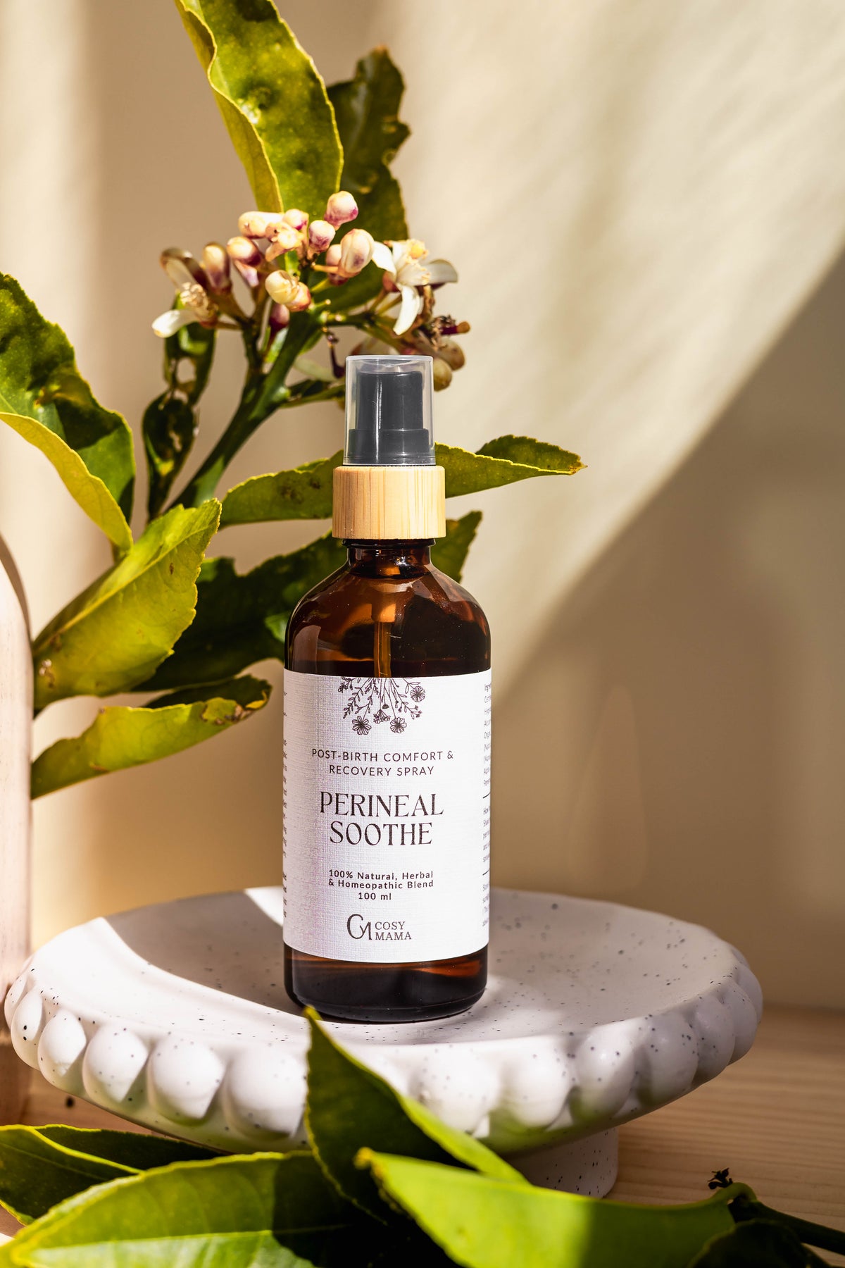A bottle of Perineal Soothe post-birth comfort and recovery spray by Cosy Mama, styled on a white ceramic pedestal with citrus leaves and small flowers under warm natural light.