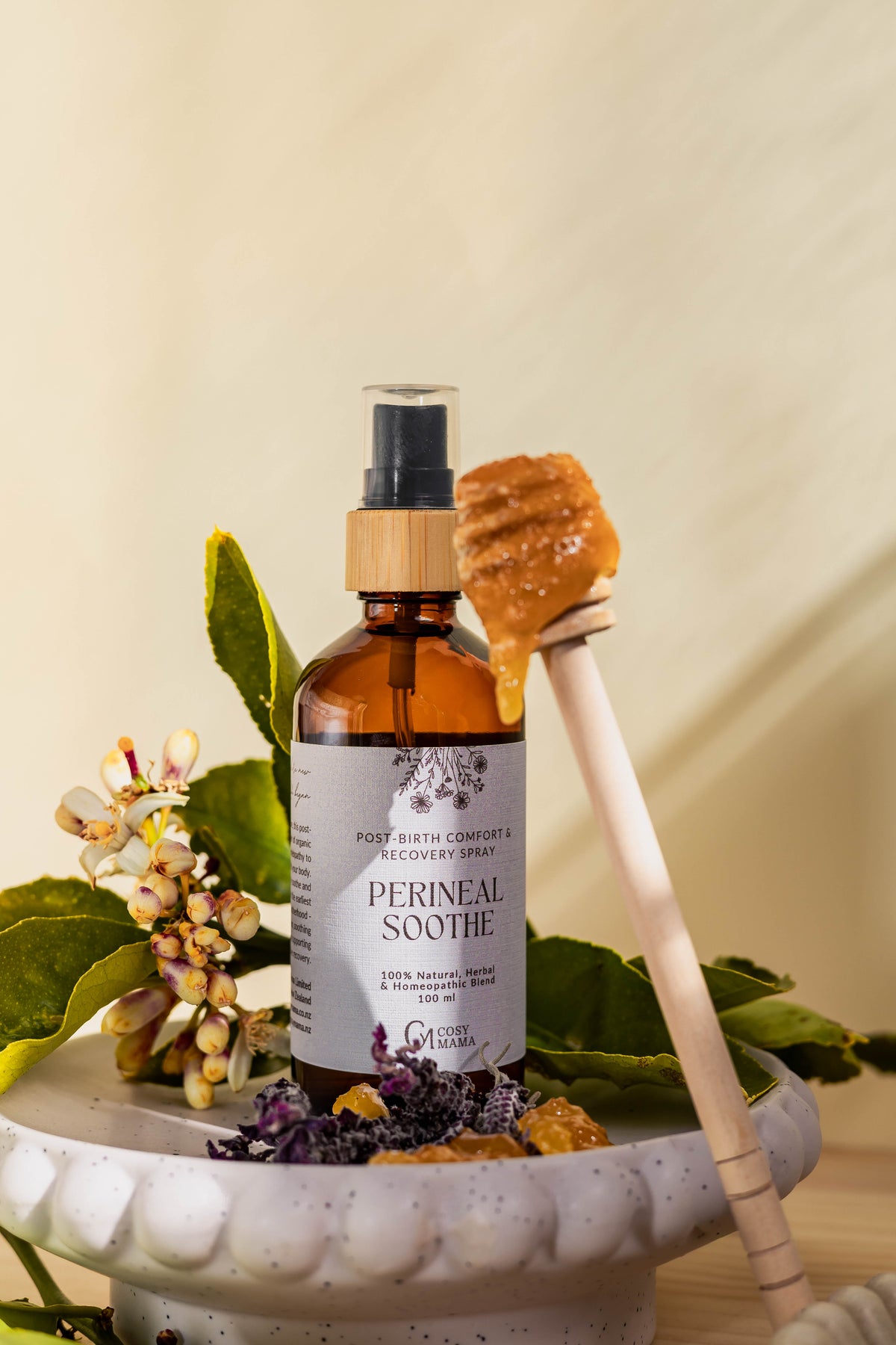 An amber glass spray bottle labeled 'PERINEAL SOOTH' stands on a white ceramic dish with botanical props, including green lemon leaves with small white flowers, Lavender, and a wooden honey dipper holding honey, against a warm cream background.