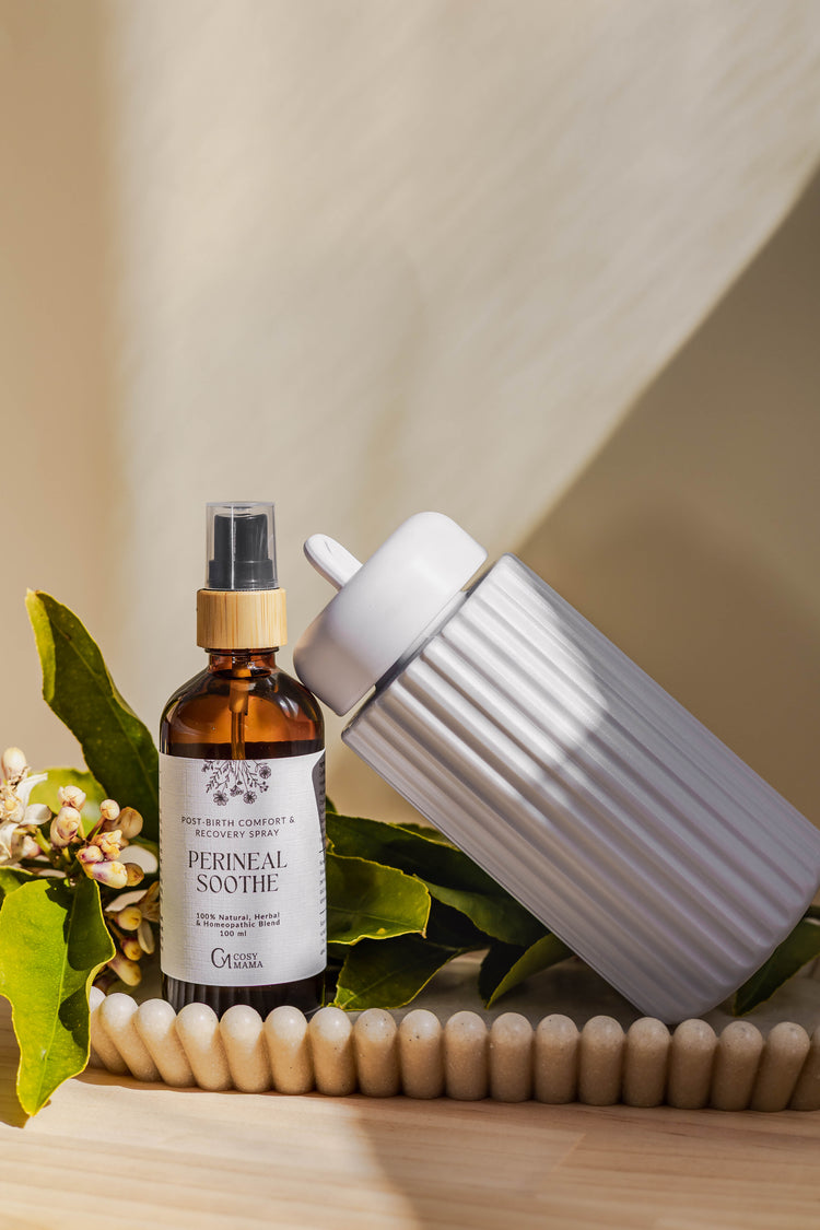 An amber glass spray bottle labeled 'PERINEAL SOOTHE' with a white label and black pump, styled next to a ribbed white Peri Spray Bottle  on a tray with green leaves and small flowers against a warm beige background.