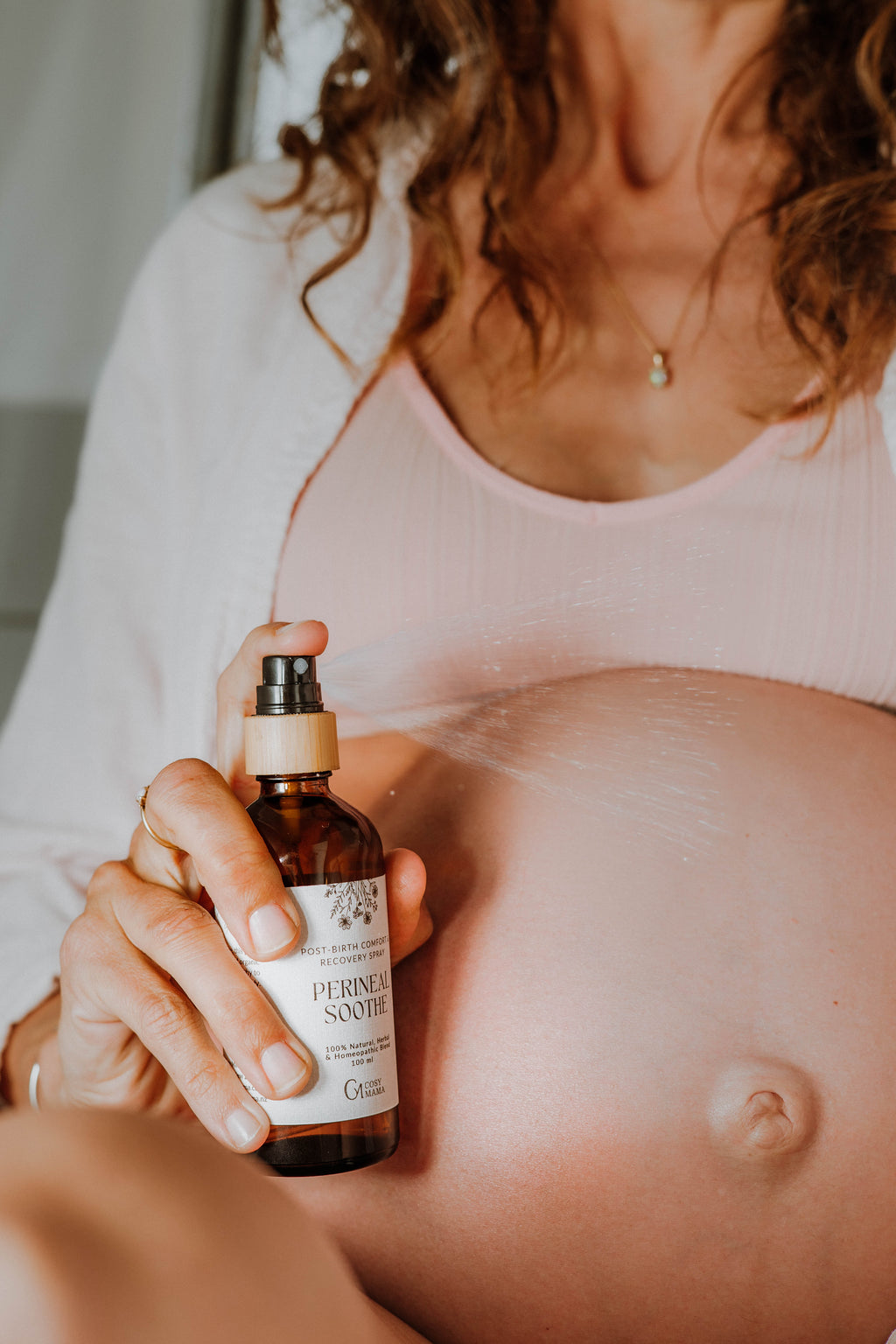 A pregnant woman holding and spraying an amber glass bottle labeled 'PerinealSoothe' onto her exposed stomach, wearing a pink ribbed top in a softly lit indoor setting.