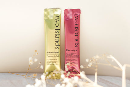 Two 'Two Islands' electrolyte powder packets on a light background with decorative dried flower elements.