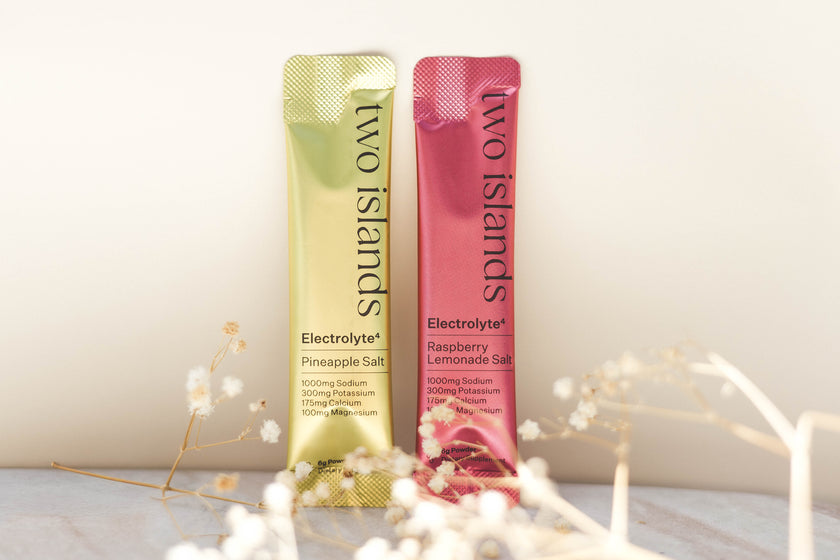 Two 'Two Islands' electrolyte powder packets on a light background with decorative dried flower elements.