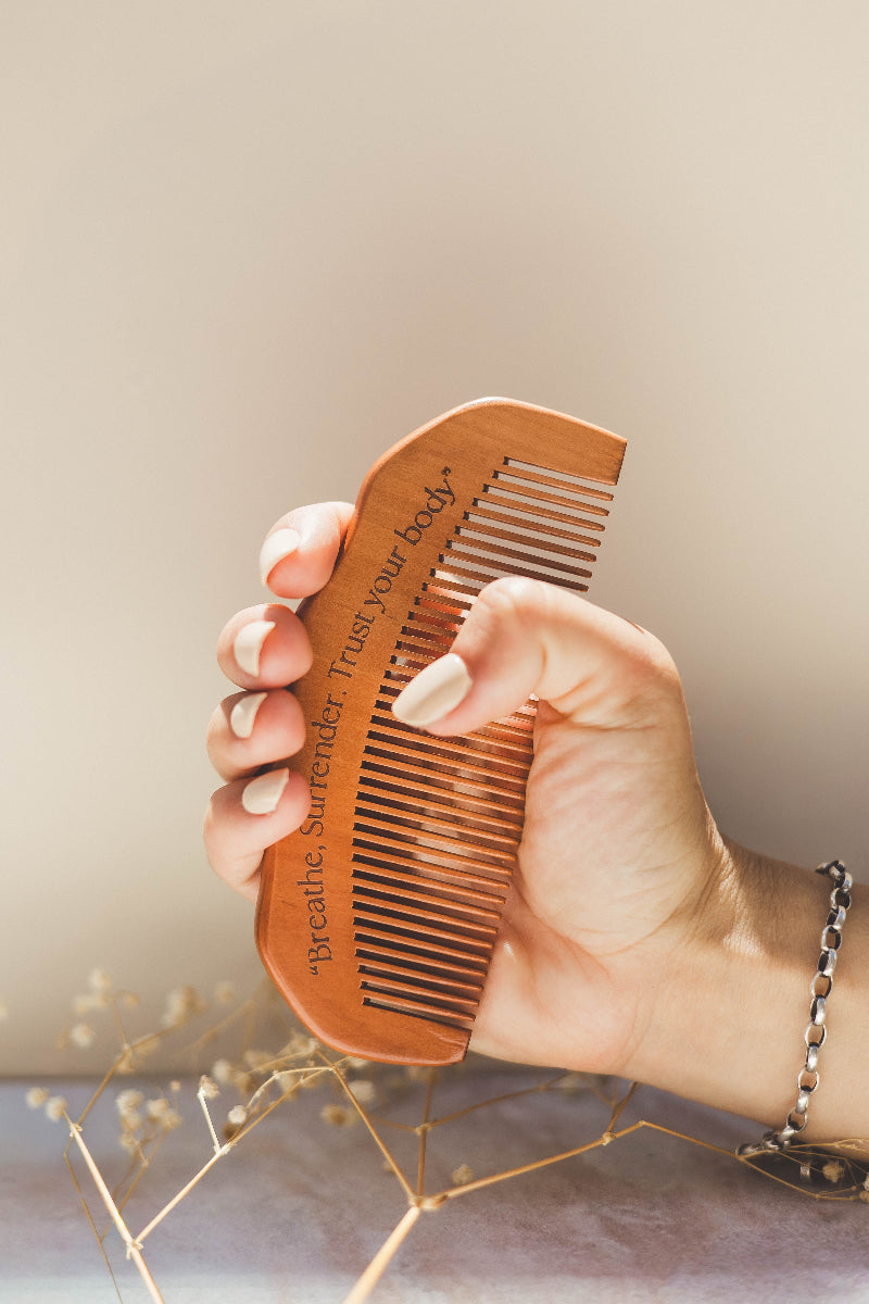 Hand holding a wooden birth comb with engraved text against a neutral background with the words 'breathe, surrender, Trust your Body'.
