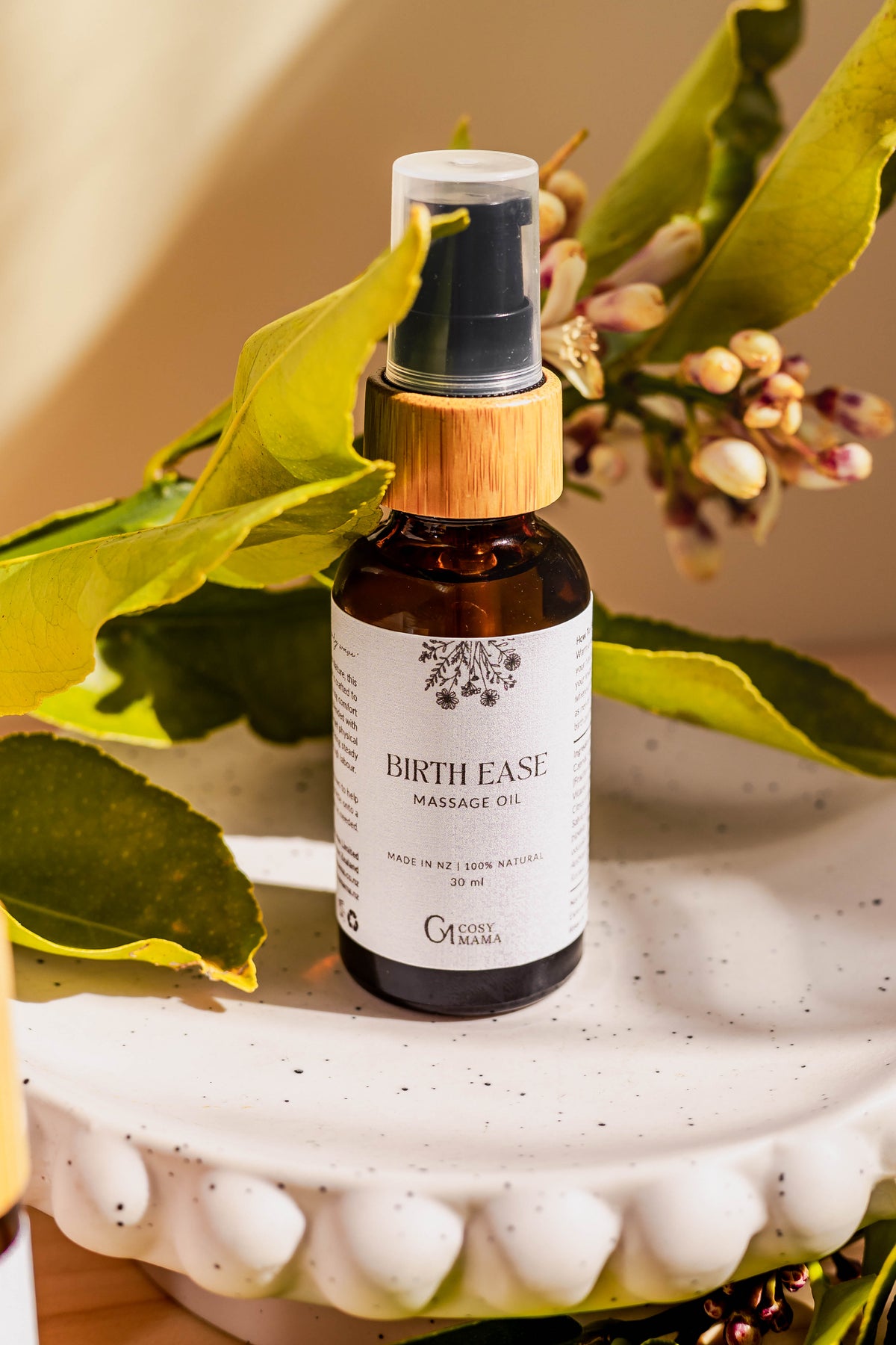 A bottle of Birth Ease Massage Oil by Cosy Mama on a white scalloped ceramic dish, surrounded by green leaves and small flowers in a warm, natural setting.