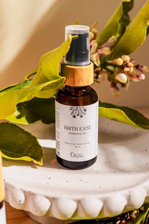 A bottle of Birth Ease Massage Oil by Cosy Mama on a white scalloped ceramic dish, surrounded by green leaves and small flowers in a warm, natural setting.